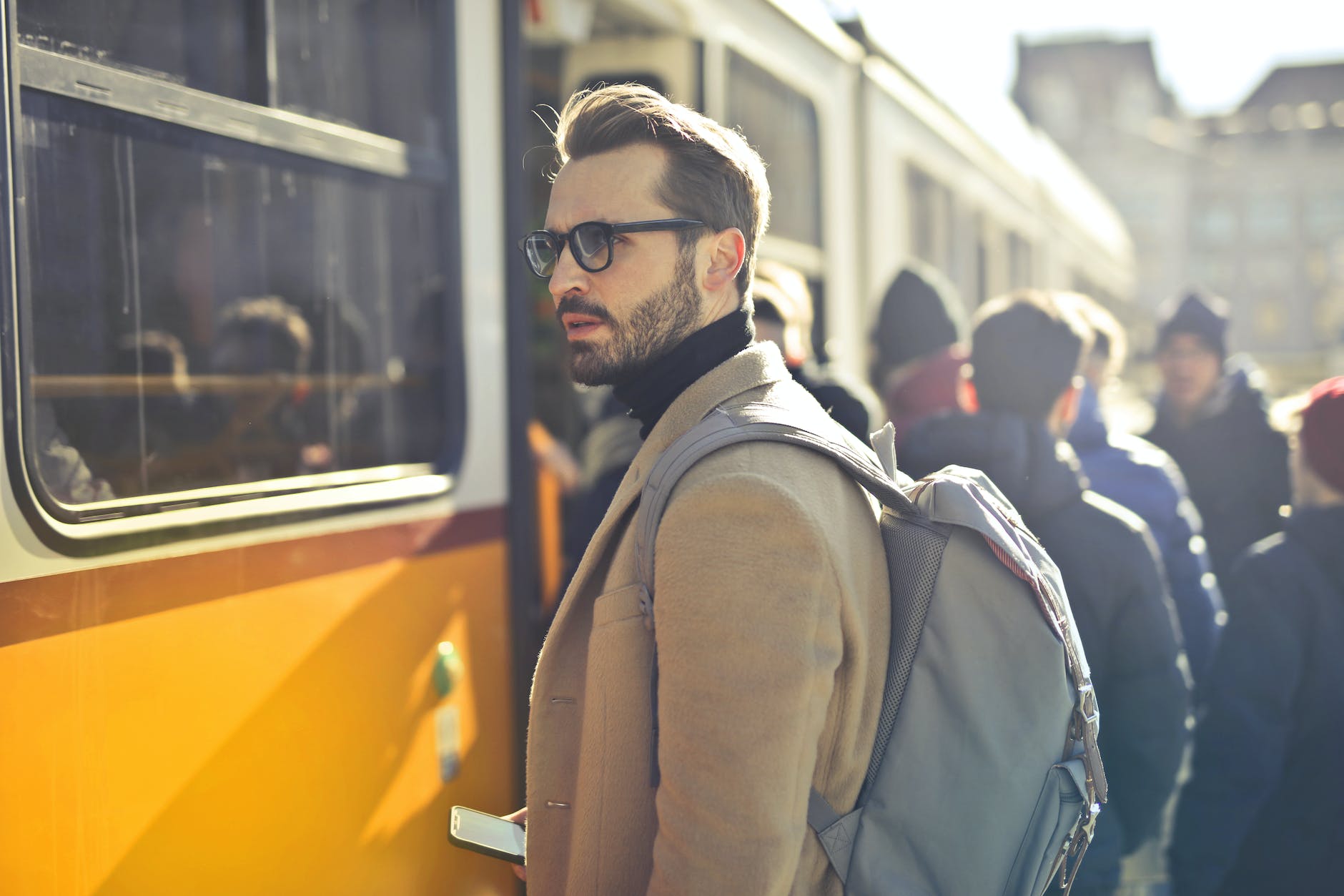man in brown coat and gray laptop backpack with cell phone near a train