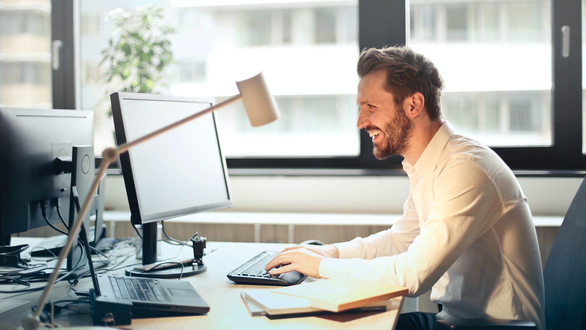 man in white dress shirt sitting on black rolling chair while facing black computer set and smiling